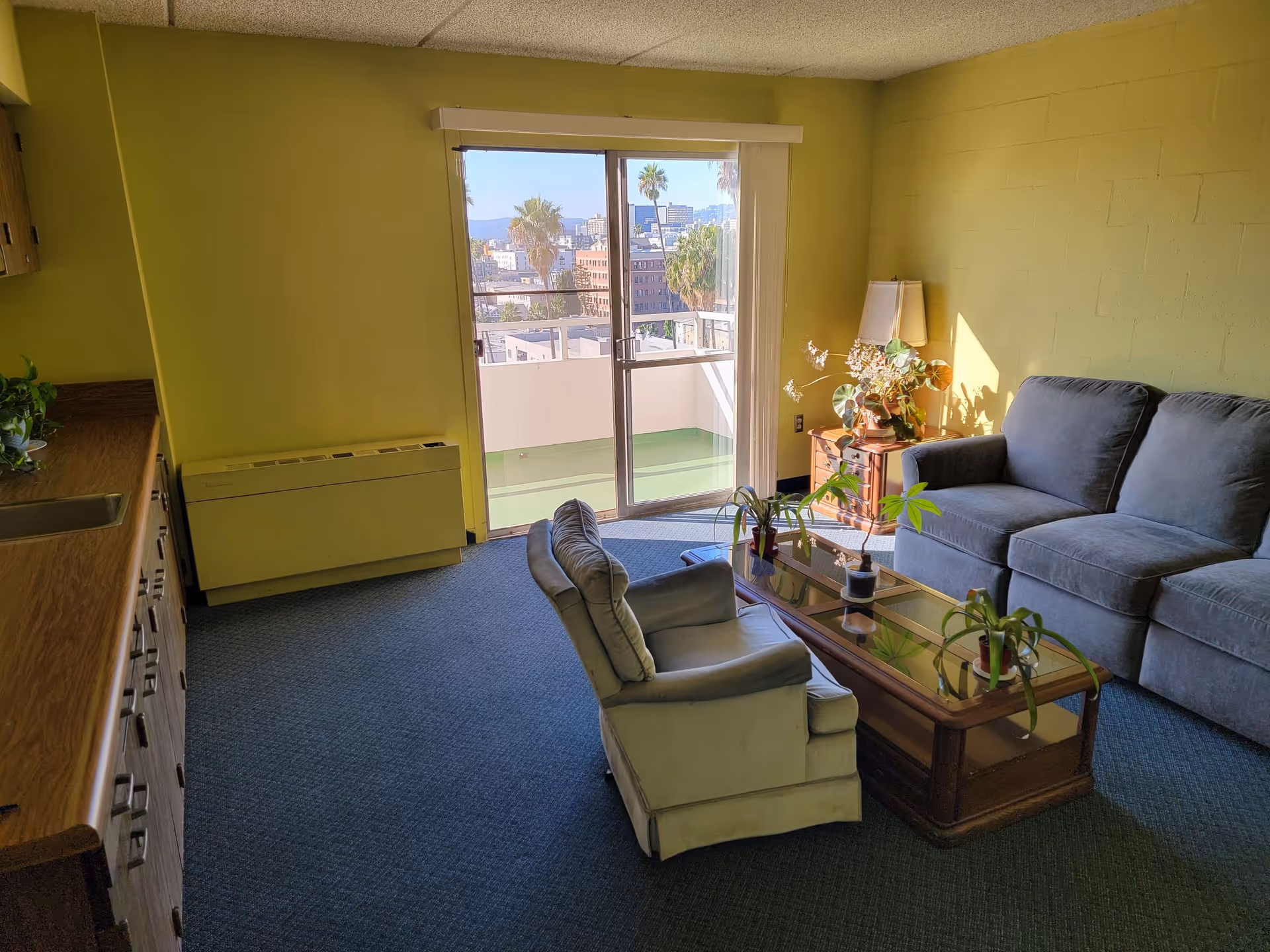 A cozy living room with a gray sofa, a cushioned armchair, and a glass coffee table with potted plants. There is a wooden side table with a lamp and decorative flowers next to the sofa. The room has yellow walls, blue carpet, and a sliding glass door leading to a balcony with a view of palm trees and city buildings.
