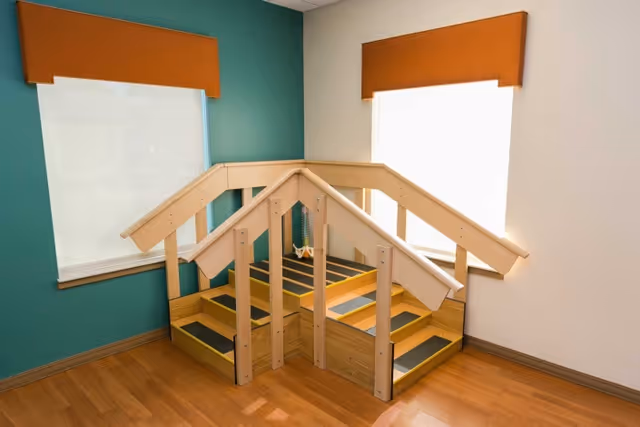Small indoor therapy staircase with wooden handrails in a corner room with teal and white walls and two windows with orange valances.