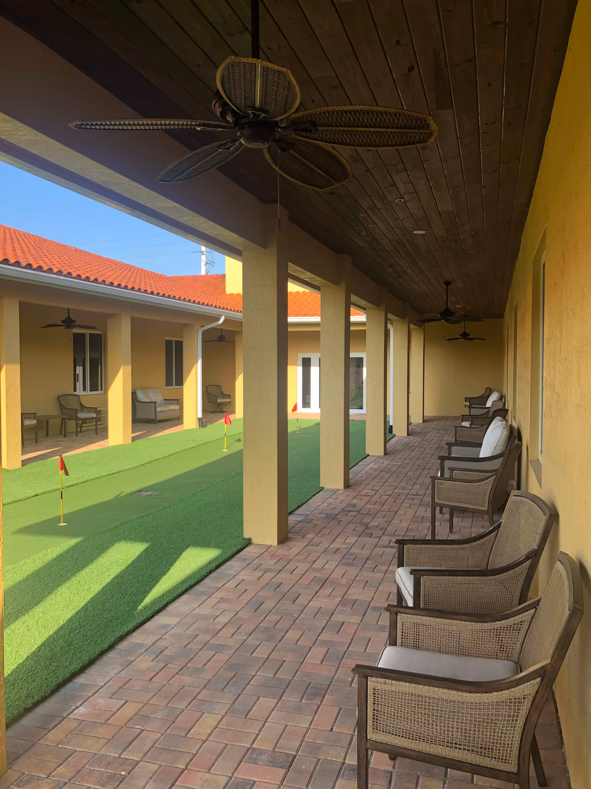 Covered outdoor patio area with wicker chairs lined up along a yellow wall and ceiling fans overhead. The patio overlooks a small artificial putting green with several red flags, surrounded by a building with a red tile roof.