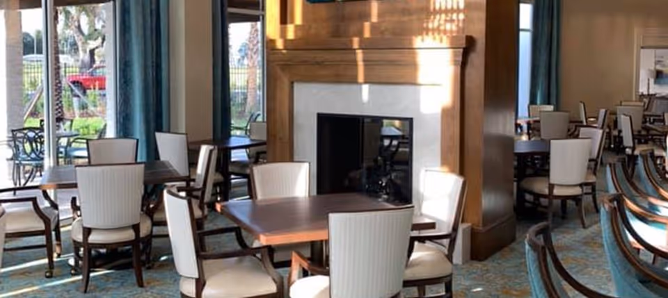 Well-lit dining room with multiple wooden tables and upholstered chairs arranged around a central fireplace and large windows.