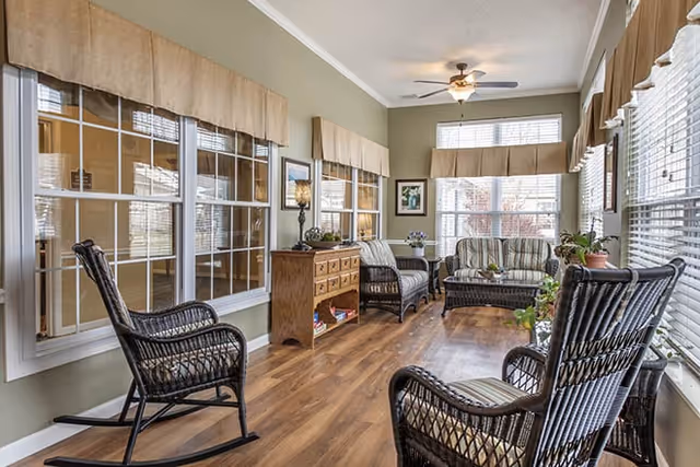 A bright and cozy sunroom with multiple large windows covered by beige valances, featuring wicker rocking chairs and a wicker loveseat with cushions, a wooden sideboard with decorative items, a ceiling fan with light, and wood flooring.