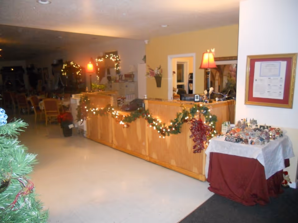Interior view of a senior living facility reception area decorated for the holidays with garlands and string lights on the wooden front desk. A table covered with a white and red cloth displays a miniature village. In the background, there are chairs and tables, a lamp on the desk, and framed documents on the wall.