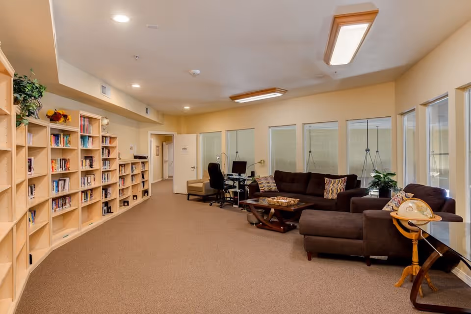 A cozy interior room in a retirement community featuring a long wooden bookshelf filled with books on the left side, a seating area with brown sofas and patterned pillows on the right, a wooden coffee table, a globe on a stand, and a computer desk with a chair near the back wall. The room has beige walls, carpeted floor, and multiple windows along the right wall.