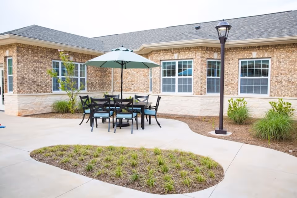 Outdoor patio area at Lavender Hills Fort Mill with a round table, six chairs with blue cushions, and a large umbrella. The patio is surrounded by a brick building with multiple windows and some landscaping including small bushes and grass patches.