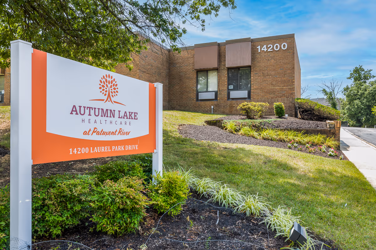 Exterior view of Autumn Lake Healthcare at Patuxent River building with a large sign displaying the facility name and address 14200 Laurel Park Drive. The building is made of brown brick with windows, surrounded by landscaped greenery and a clear blue sky.
