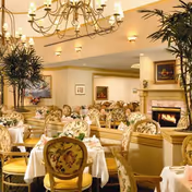 Elegant dining room with multiple tables set with white tablecloths, floral-upholstered chairs, chandeliers, and a fireplace.