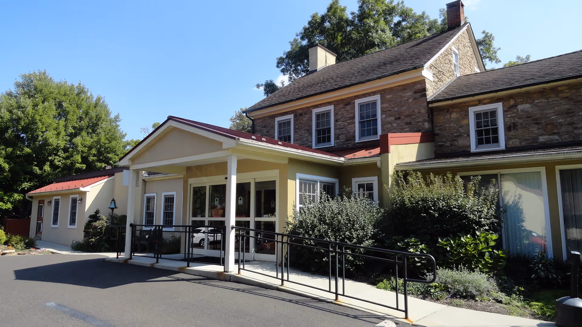 Exterior view of a senior living facility building with a stone and yellow facade, featuring a covered entrance with a ramp and handrails, surrounded by greenery and trees under a clear blue sky.