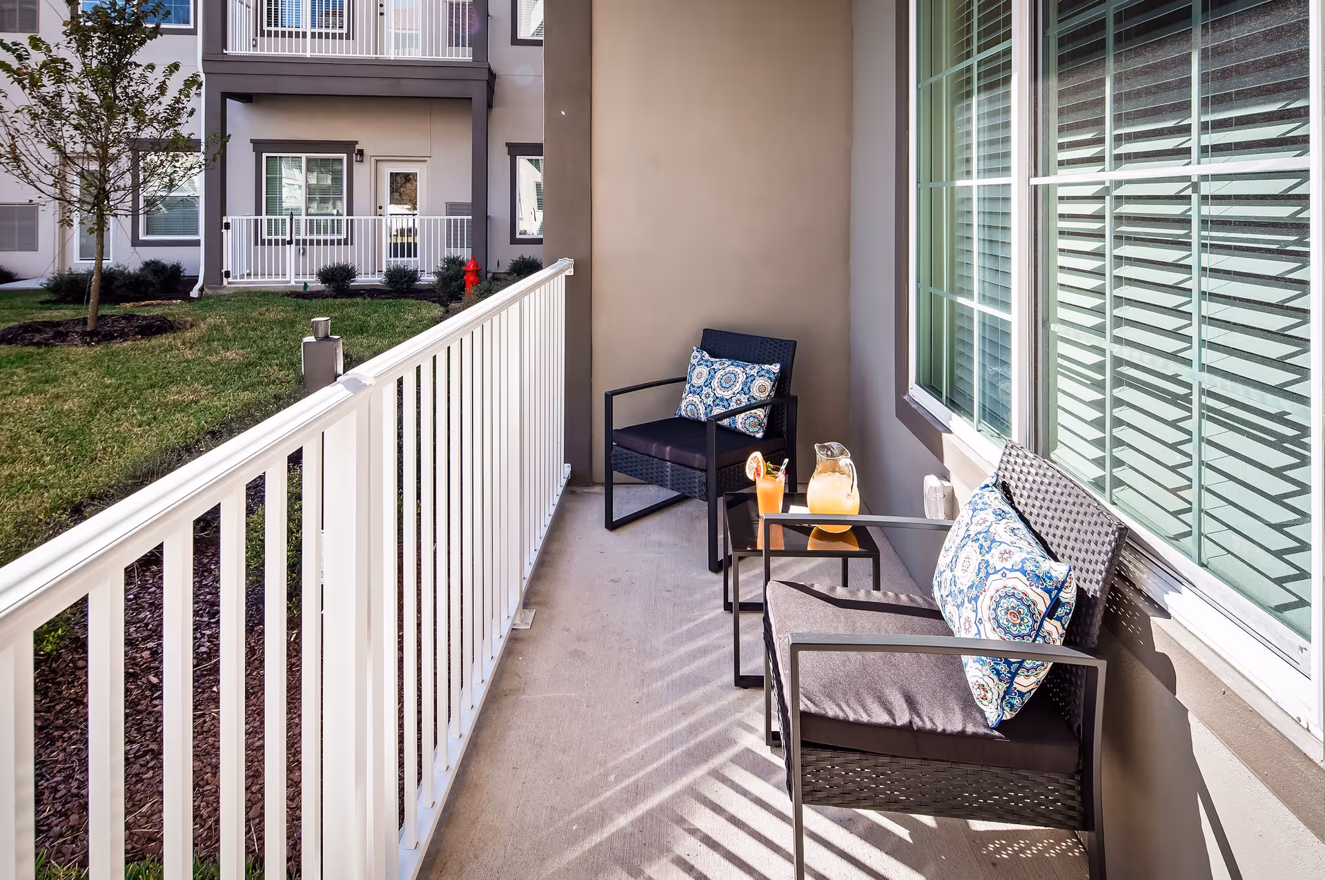 A small outdoor patio area with two black wicker chairs with patterned cushions and a small black table between them holding a pitcher of lemonade and a glass with a straw and garnish. The patio is enclosed by a white railing and overlooks a grassy area with a tree and nearby building windows and doors.