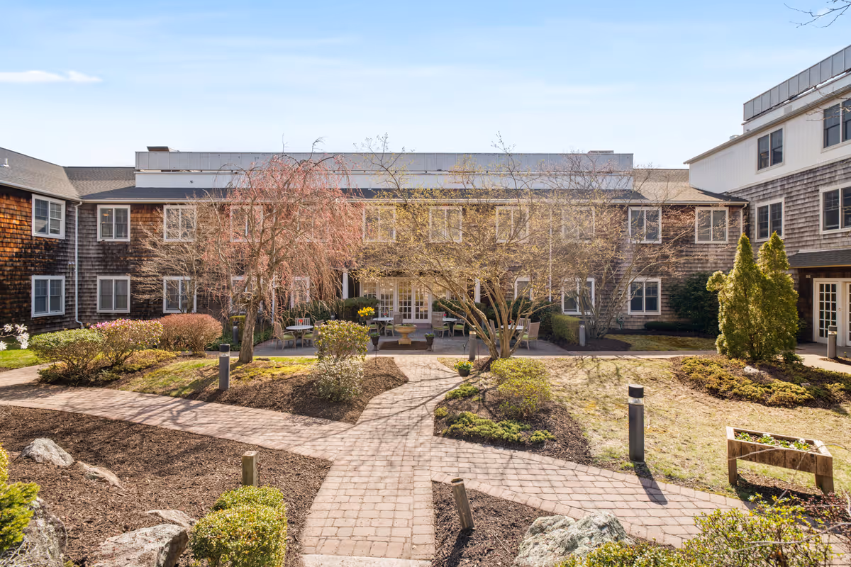 Outdoor courtyard area of a senior living facility with paved walkways, landscaped garden beds, small trees, shrubs, and outdoor seating. The building surrounding the courtyard has multiple windows and a rustic exterior with wooden shingles.