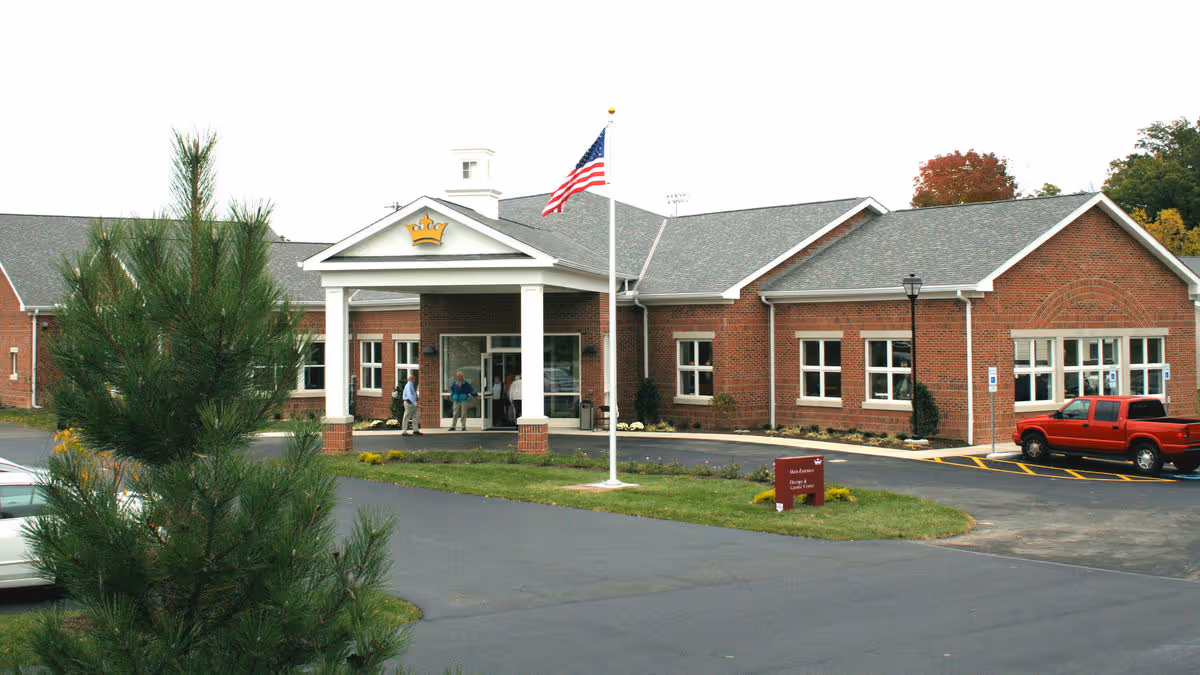 Brick single-story senior living facility entrance with a covered portico, American flag, parked cars, and people near the doorway.