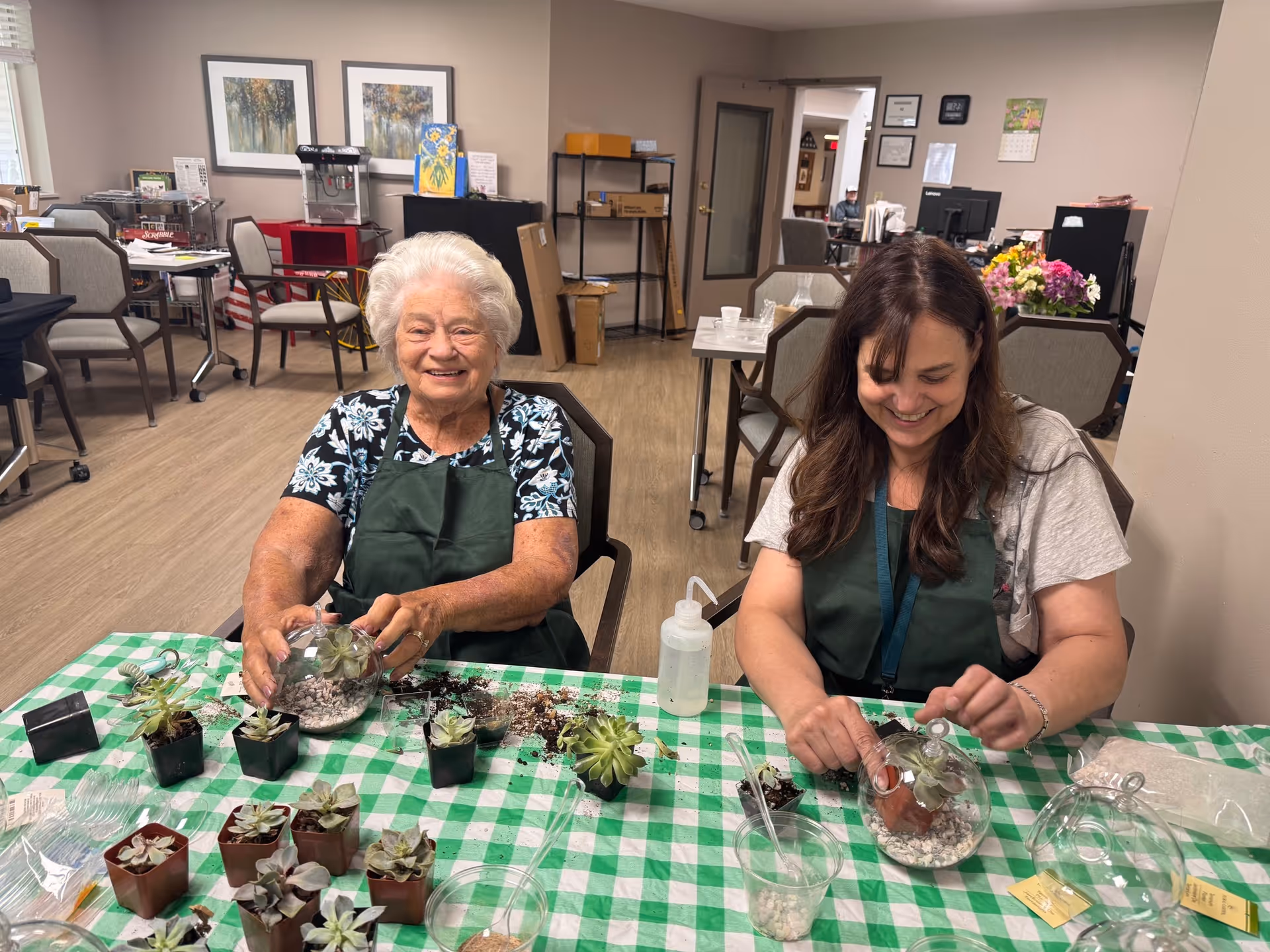 Two women sitting at a table covered with a green and white checkered tablecloth, working on planting small succulents in glass containers. The older woman on the left is smiling at the camera, while the younger woman on the right is focused on her planting. The room has chairs, tables, and office equipment in the background.