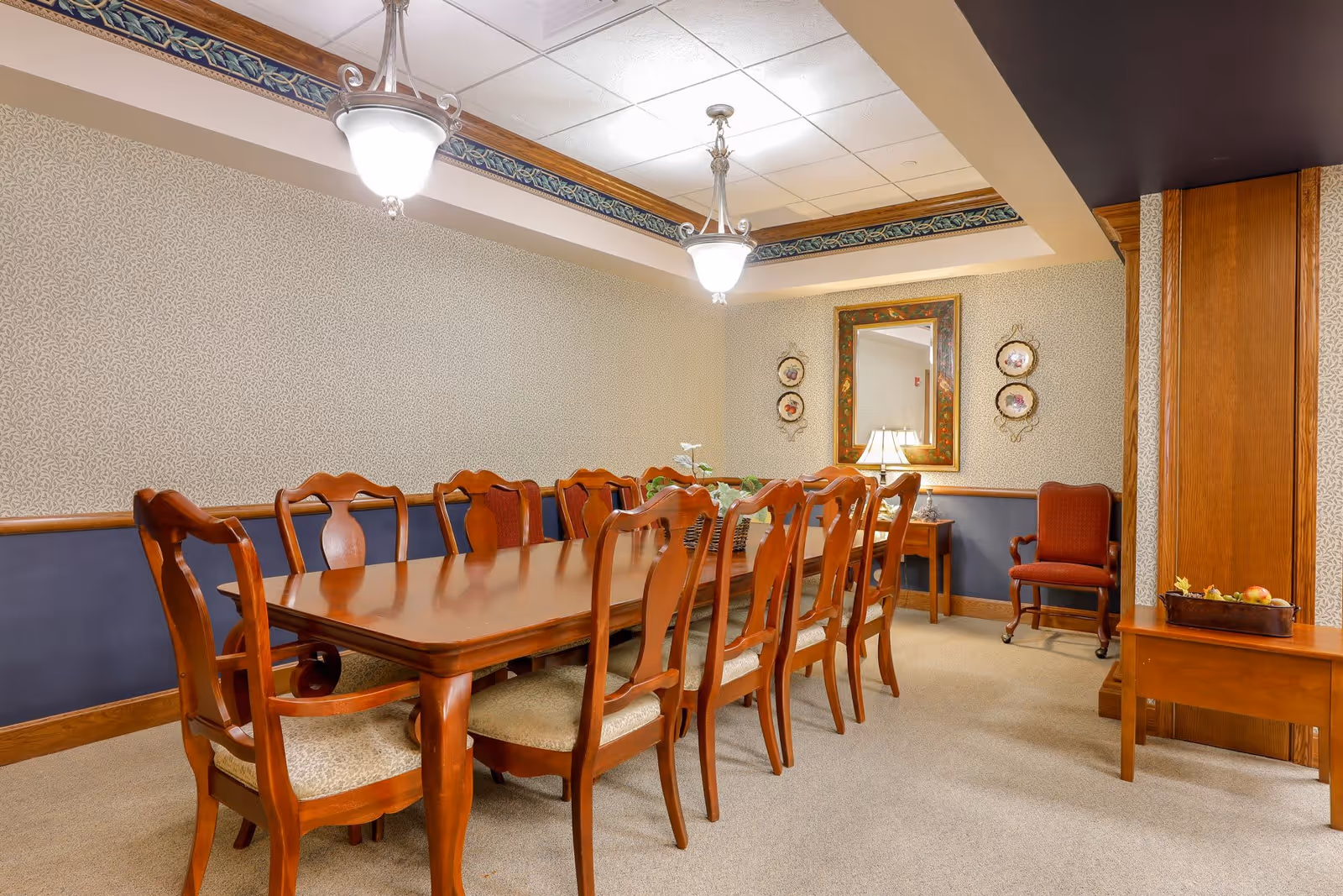 A formal dining room with a long wooden table surrounded by ten wooden chairs with cushioned seats. The room has patterned wallpaper with a blue wainscot, two hanging light fixtures, a framed mirror on the wall, decorative plates, a table lamp on a side table, and an upholstered armchair in the corner.