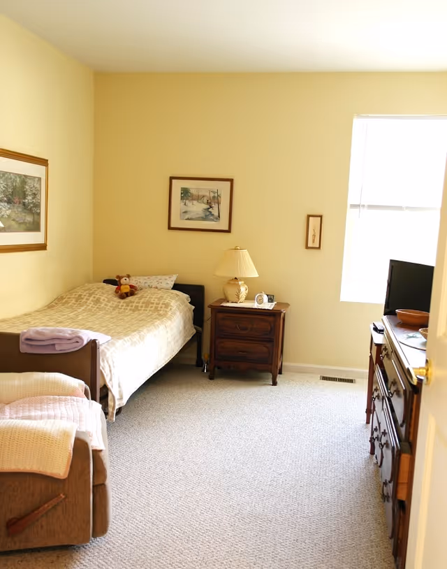 A cozy bedroom with a single bed covered in a beige patterned blanket and a teddy bear on the pillow. Next to the bed is a wooden nightstand with a lamp, clock, and small items. The room has light yellow walls with framed artwork, a window letting in natural light, a wooden dresser with a TV on top, and a beige carpeted floor.