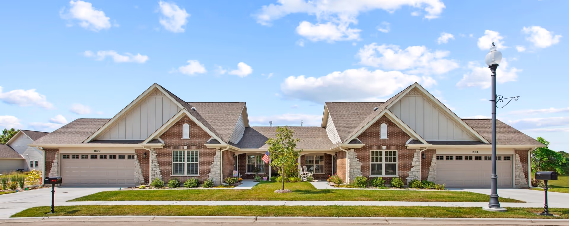 Exterior view of a single-story residential building with two attached garages, brick and siding facade, a central entrance with a small porch, manicured lawn, and a streetlamp on the sidewalk under a partly cloudy sky.