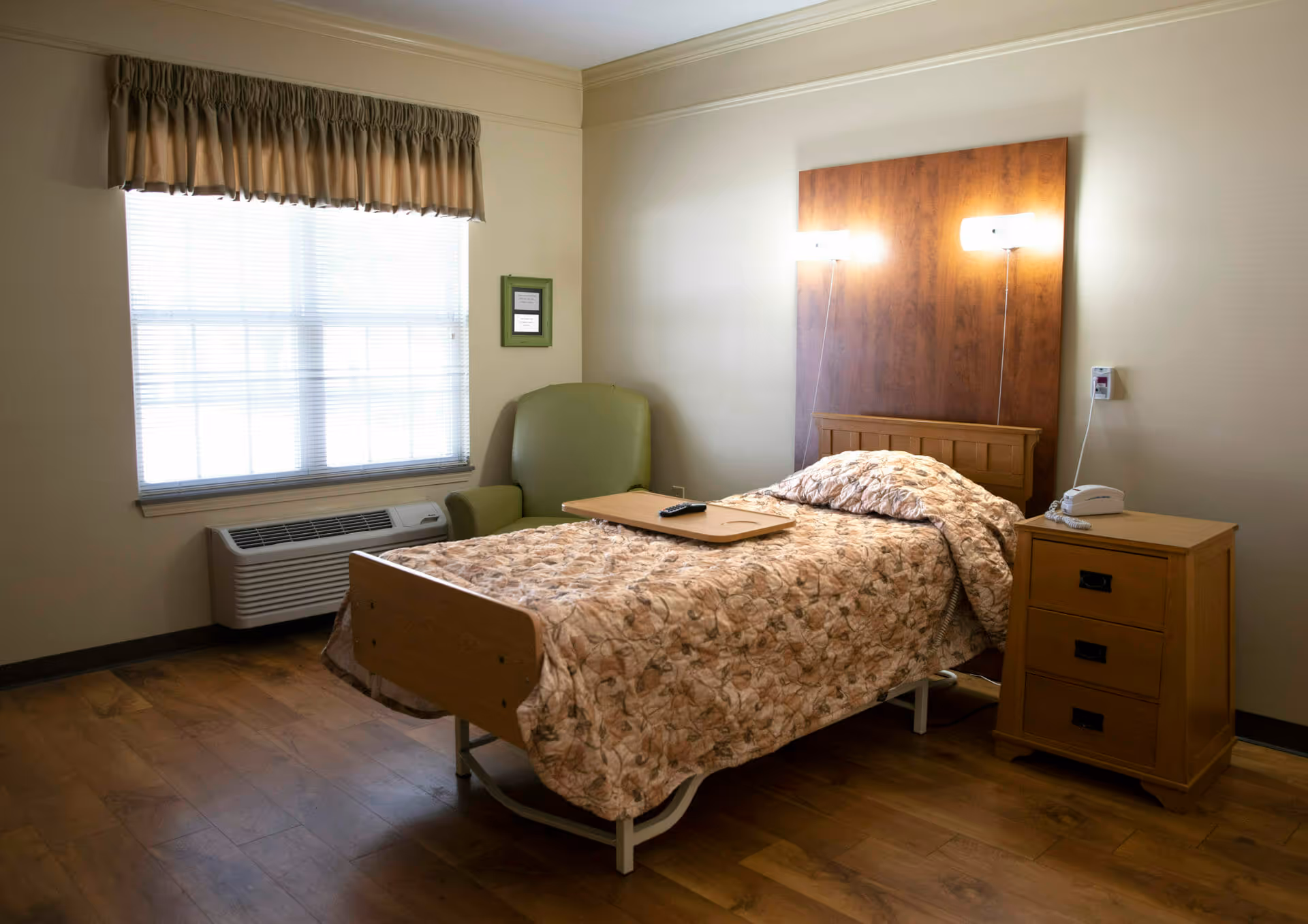 A simple furnished single-occupancy bedroom with a bed, bedside table, armchair, and a window with a valance.