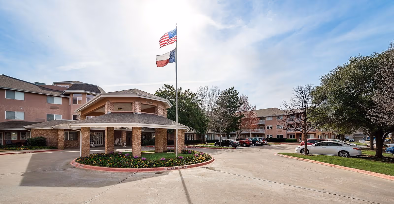Exterior view of The Bentley - Sky Active Living facility showing a covered entrance with brick pillars, a circular driveway, landscaped flower beds, and two flagpoles flying the American and Texas flags. Several parked cars and trees surround the building under a partly cloudy sky.