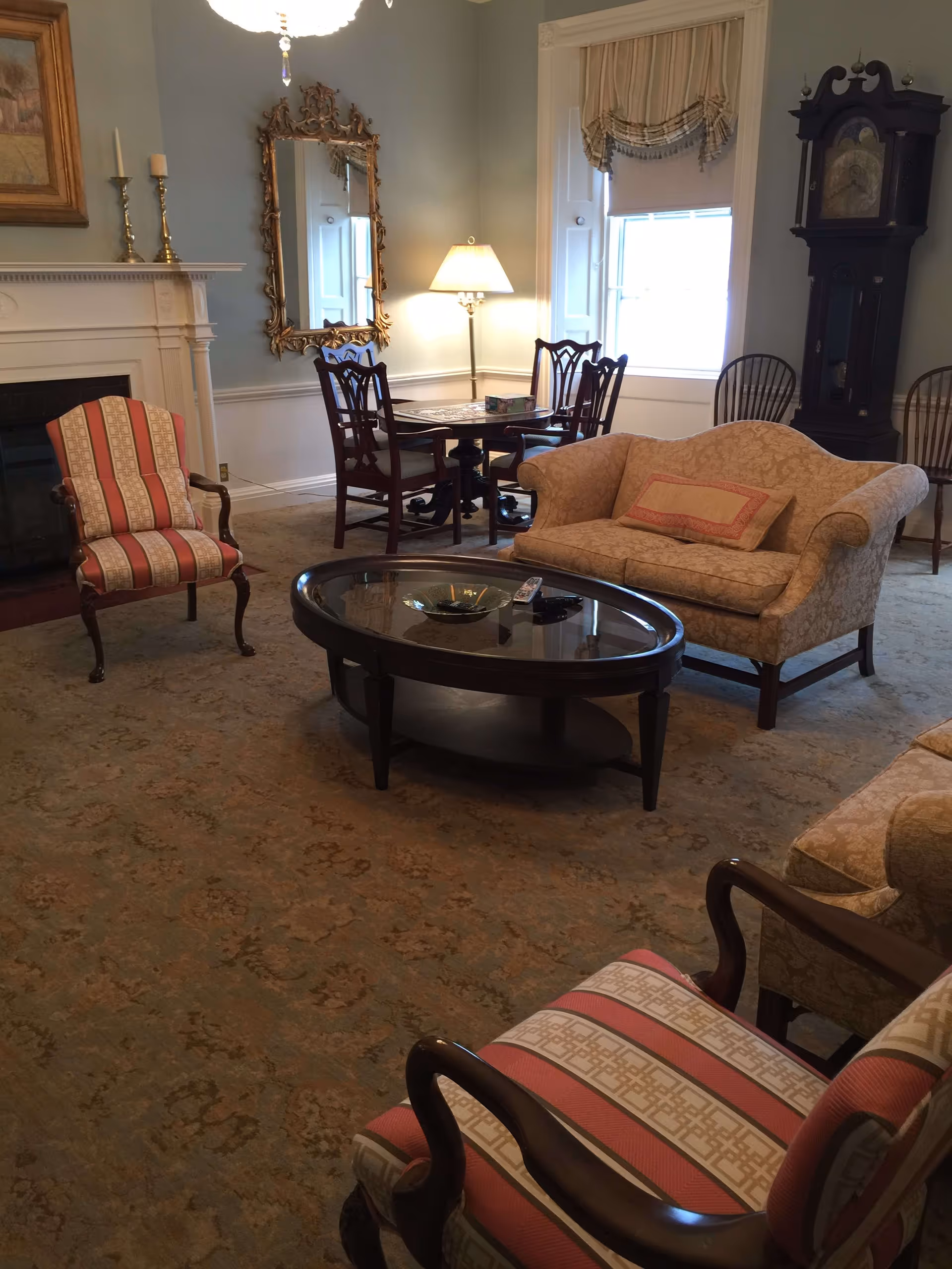 A cozy living room with classic furniture including a beige patterned loveseat with a decorative pillow, two striped armchairs, a dark wood oval coffee table with a glass top, and a round dining table with four chairs near a window. The room features a fireplace with candlesticks on the mantel, a tall grandfather clock, a decorative mirror on the wall, and a floor lamp providing warm lighting.