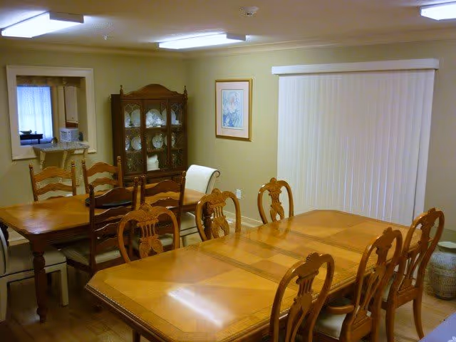 A dining room with two wooden dining tables and several matching wooden chairs. There is a china cabinet with glass doors displaying dishes against the far wall. A framed picture hangs on the wall next to vertical blinds covering a window. The room has beige walls and a ceiling light fixture.