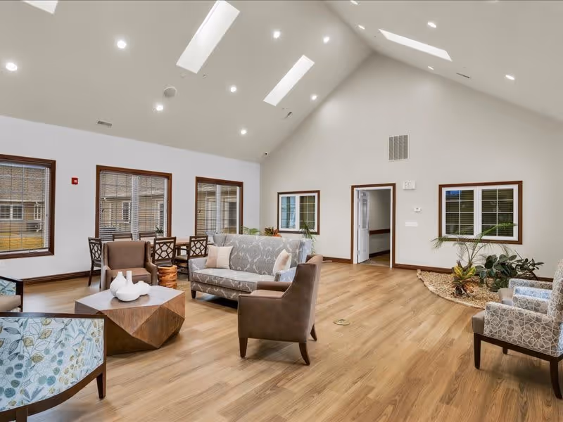 A spacious and bright living room area with high vaulted ceilings and skylights. The room features a mix of patterned and solid armchairs, a patterned sofa, a wooden geometric coffee table with decorative white vases, and a dining table with chairs near three large windows. There is a small indoor garden with plants in the corner and light wood flooring throughout.