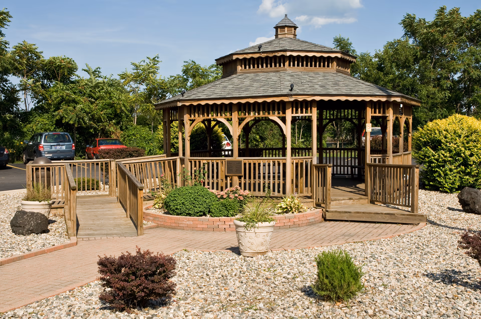 A wooden gazebo with a shingled roof surrounded by a landscaped garden with bushes, plants, and a stone-covered ground. There are ramps leading up to the gazebo, and cars are parked in the background under a clear blue sky.