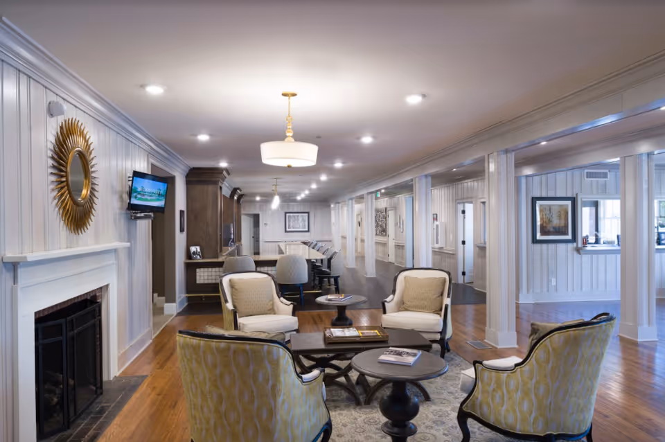 A spacious and well-lit common area with comfortable seating including armchairs and small tables arranged on a rug. The room features hardwood floors, white paneled walls, a fireplace with a decorative sunburst mirror above it, and a wall-mounted TV. In the background, there is a long counter with bar stools and additional seating, with framed artwork on the walls and recessed lighting on the ceiling.