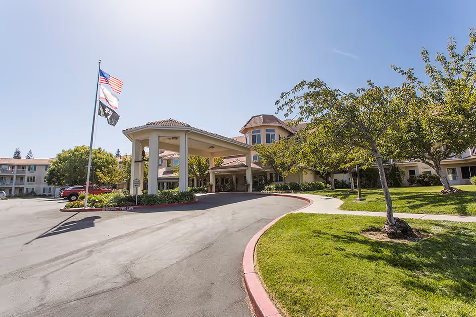 Exterior view of Quail Lodge Retirement Community showing the main entrance with a covered drop-off area, a flagpole with three flags, a curved driveway, green lawns, and trees under a clear blue sky.