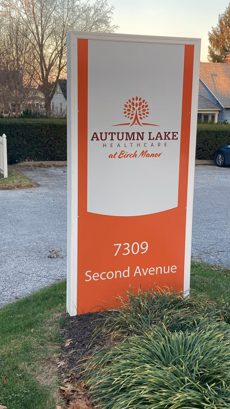 Outdoor sign for Autumn Lake Healthcare at Birch Manor located at 7309 Second Avenue, with grass and shrubs in the foreground and houses and trees in the background.