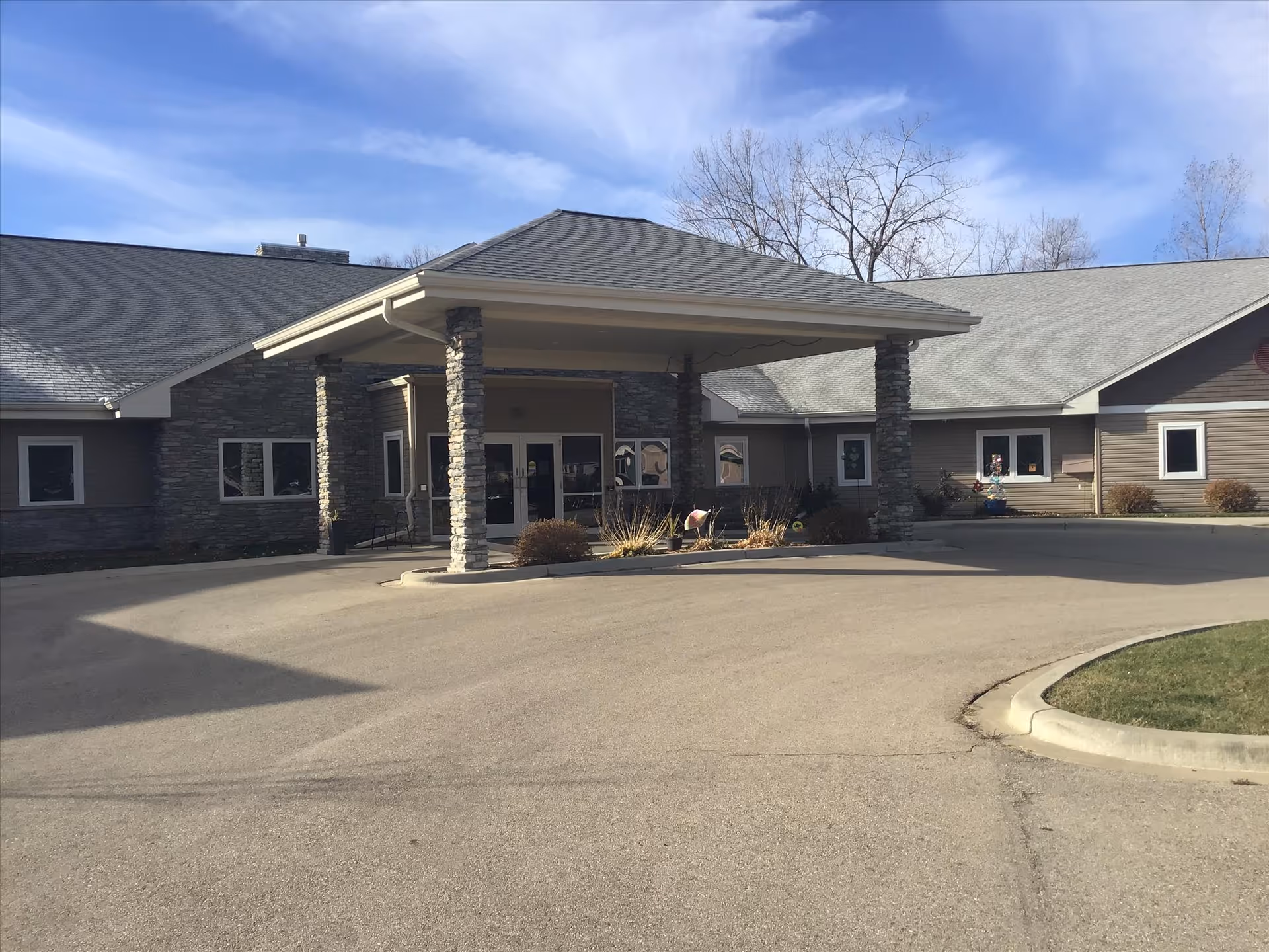 Front exterior view of Prairie Ridge Assisted Living facility showing a covered entrance supported by stone pillars, surrounded by a paved driveway and some landscaping with bare trees in the background under a partly cloudy sky.