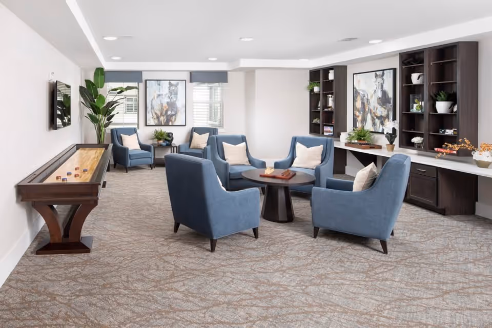A bright and spacious lounge area with six blue upholstered armchairs arranged around a circular wooden table. The room features a shuffleboard table along the left wall, large windows with blinds, two framed horse paintings, built-in dark wood shelves with decorative plants and items, and a light patterned carpet.