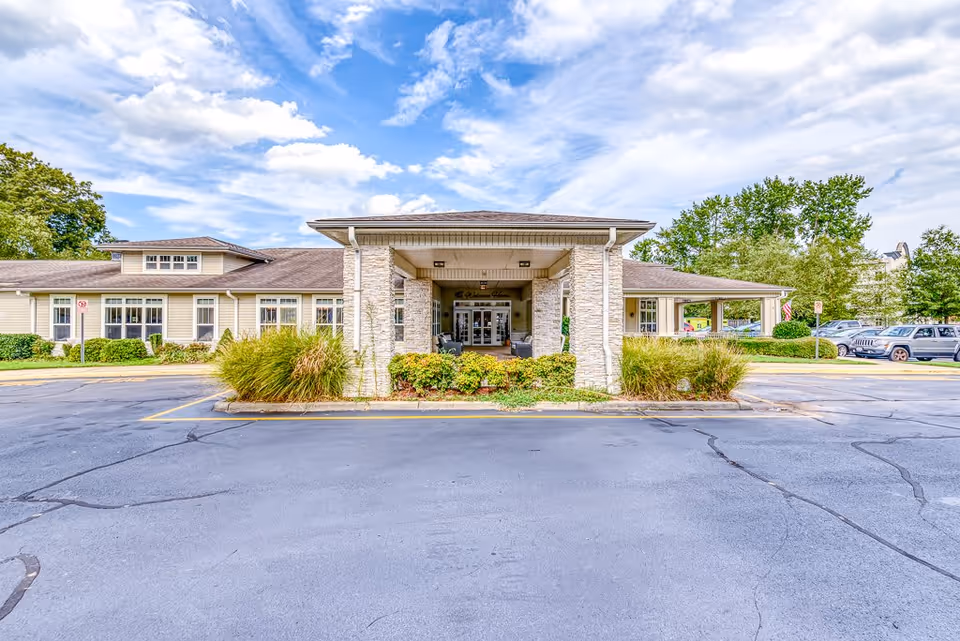 Front exterior view of Chesapeake Place Senior Living building with a covered entrance, stone pillars, surrounding greenery, parked cars, and a partly cloudy sky.