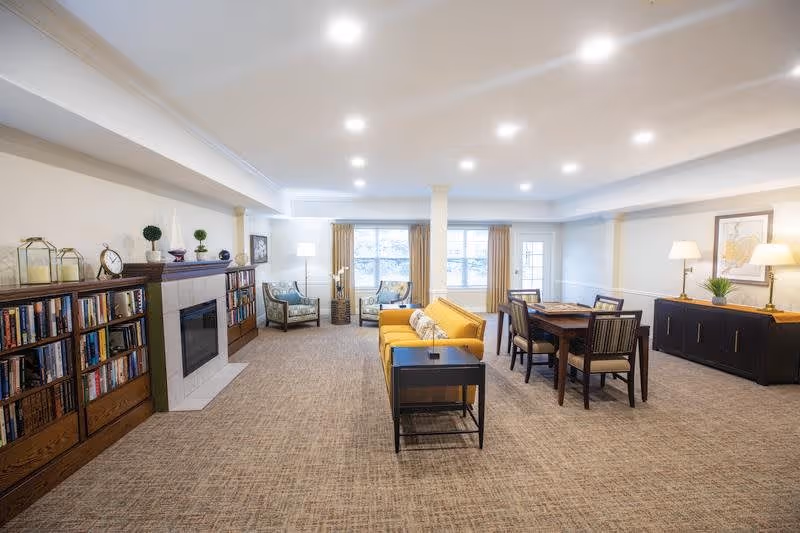 A spacious and well-lit common area in a senior living facility featuring a yellow sofa, two armchairs near large windows with beige curtains, a wooden dining table with four chairs, a fireplace flanked by bookshelves filled with books, and a sideboard with two lamps and a framed picture above it.