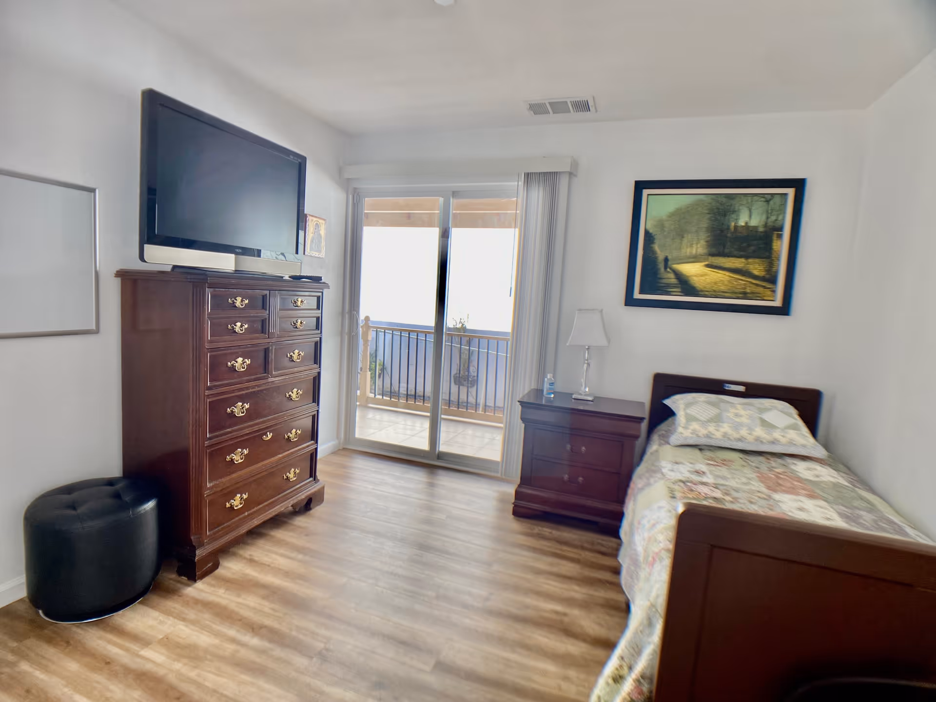 A bedroom with a single bed covered in a patchwork quilt, a wooden nightstand with a lamp and a water bottle, a tall wooden dresser with a flat-screen TV on top, a black ottoman, and a sliding glass door leading to a balcony. A framed painting hangs on the wall above the bed.