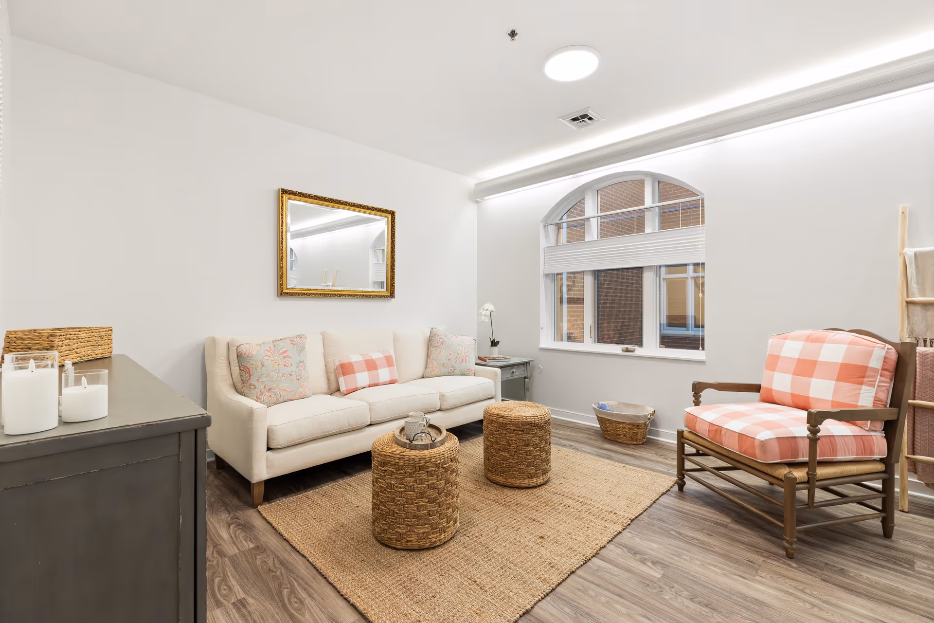 A cozy living room with a beige sofa adorned with patterned and checkered pillows, a wooden armchair with pink and white checkered cushions, two woven round ottomans on a natural fiber rug, a gray sideboard with candles, a small side table with a white orchid, and a large window with an arched top and white blinds letting in natural light.