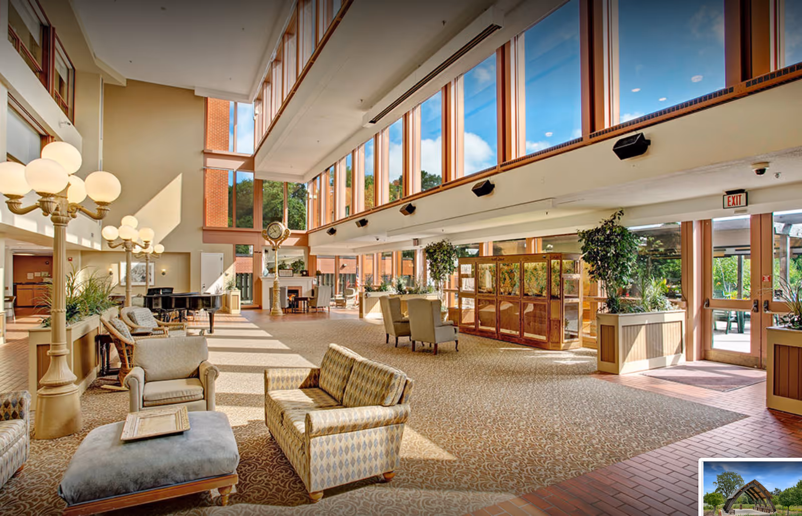 Spacious sunlit lobby with high windows, seating areas, plants, and a grand piano.