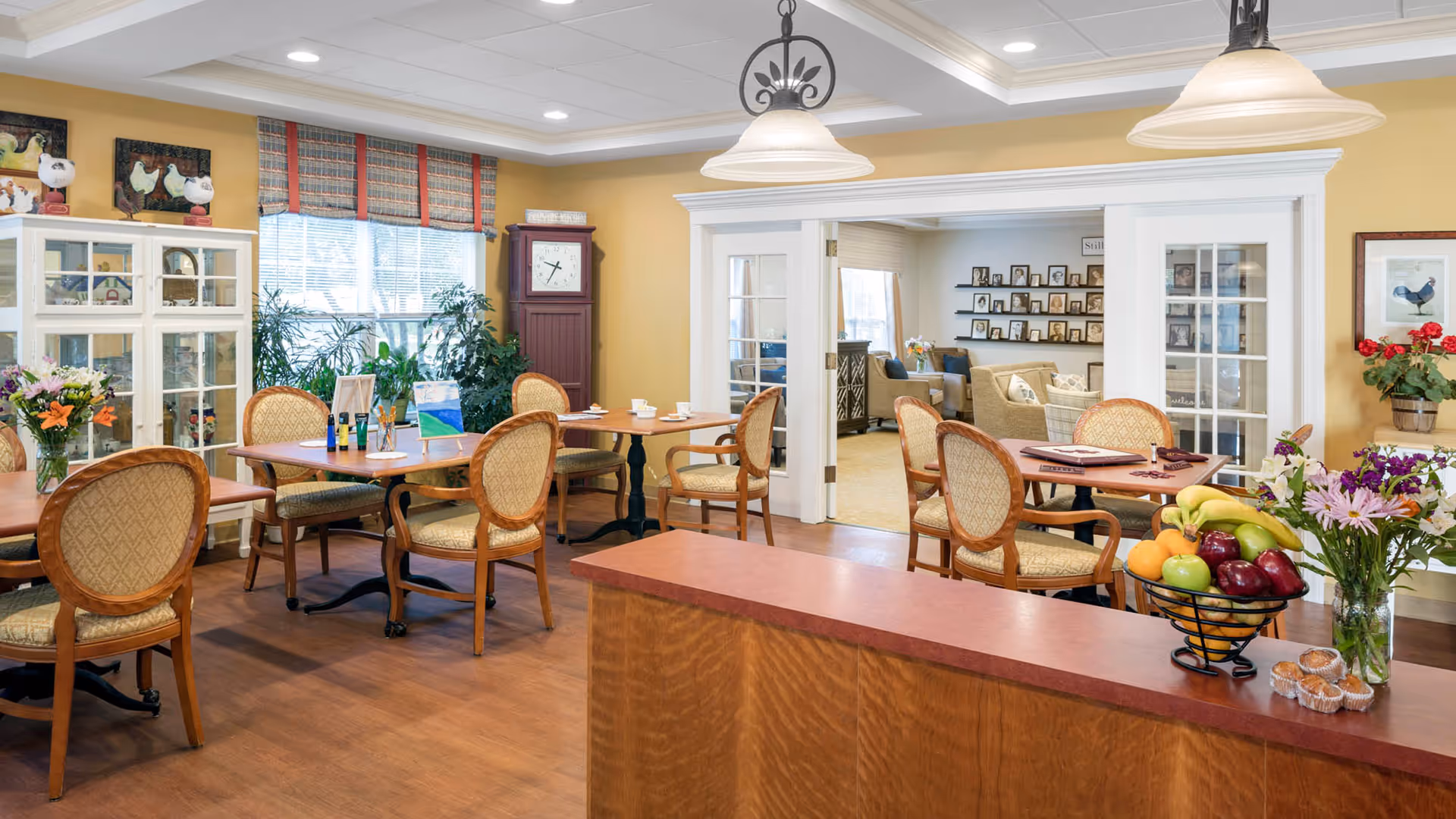A bright and inviting dining room in a senior living facility with several wooden tables and cushioned chairs. The room features a large window with plaid curtains, a tall grandfather clock, and a white display cabinet with decorative items. There are flowers in vases on the tables and a fruit basket with assorted fruits on a counter in the foreground. Through a wide doorway with glass-paneled double doors, a cozy living room with beige sofas and framed photos on shelves is visible.