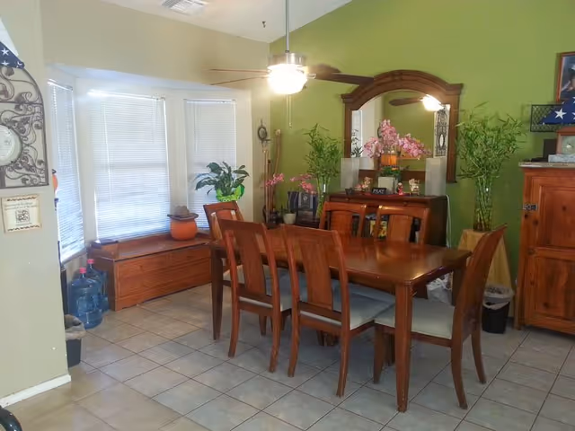 Dining room with a wooden table and six chairs, green accent wall, mirror, plants and a ceiling fan.
