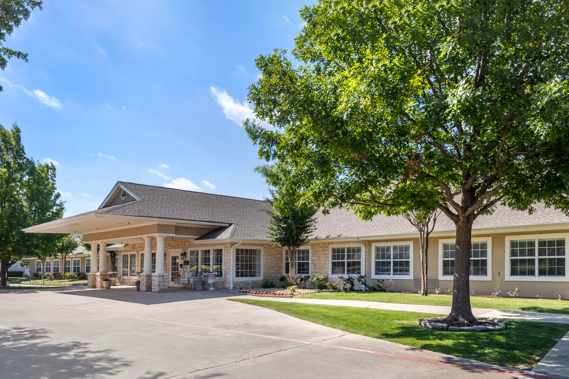 Exterior view of a single-story senior living facility building with a covered entrance supported by white columns. The building has beige walls with stone accents and multiple windows. There are green trees and well-maintained grass around the building under a clear blue sky.