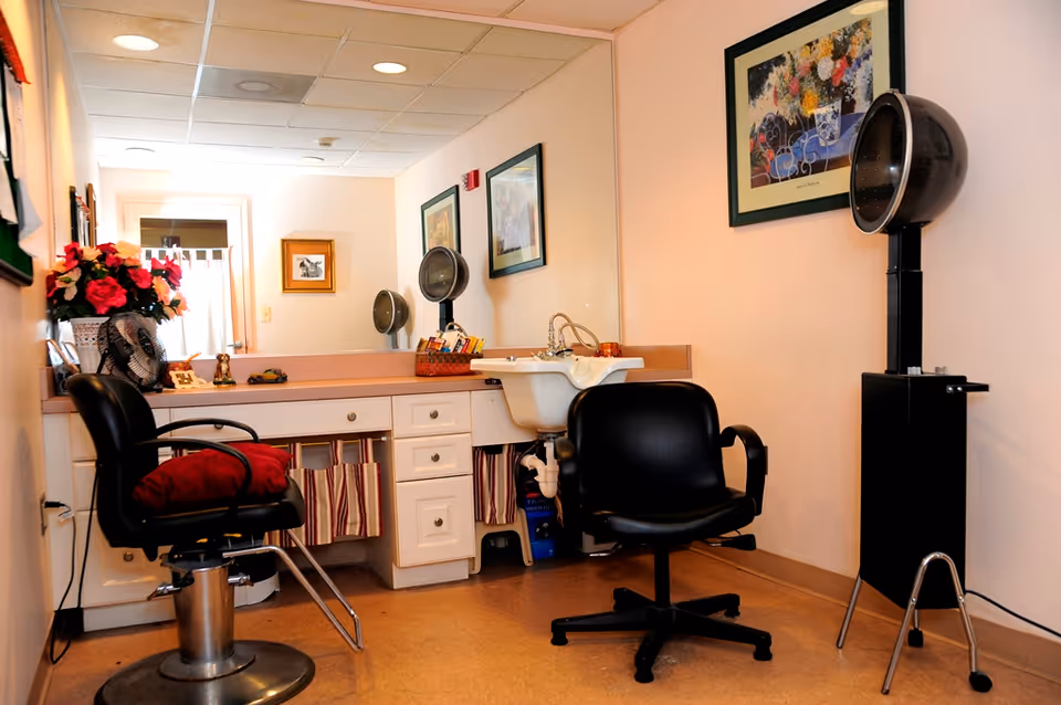 Interior view of a hair salon area in a senior living community, featuring two black salon chairs, a hair washing sink, a hair dryer hood, a large mirror, and decorative framed artwork on the walls. There is also a countertop with drawers and a vase of flowers.