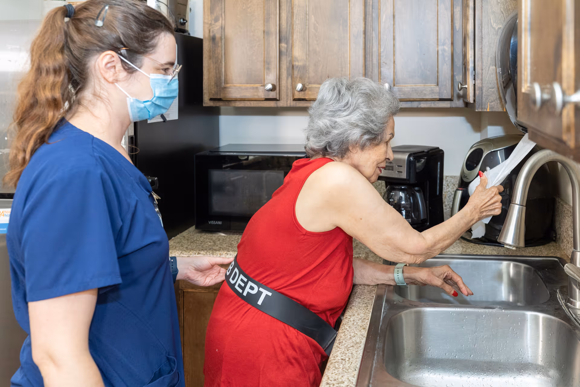 An elderly woman in a red sleeveless top is standing at a kitchen sink, pulling a paper towel from a dispenser above the sink. A caregiver wearing blue scrubs and a face mask is standing beside her, providing support by holding her back.