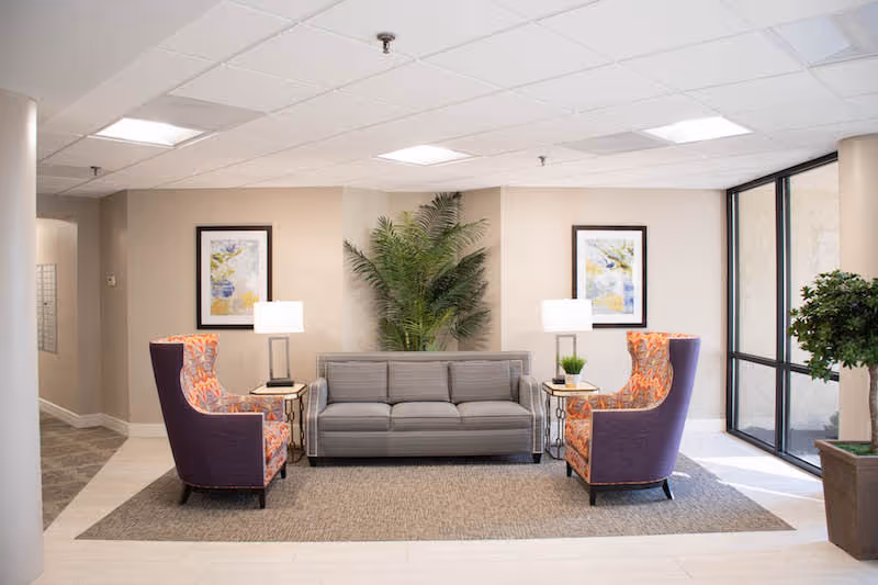 A bright and welcoming senior living community lounge area featuring a gray sofa centered between two patterned armchairs with purple backs. Two side tables with lamps and small plants flank the sofa. A large green plant is placed in the corner behind the sofa, and two framed artworks hang on the beige walls. Large windows on the right side allow natural light to fill the space.