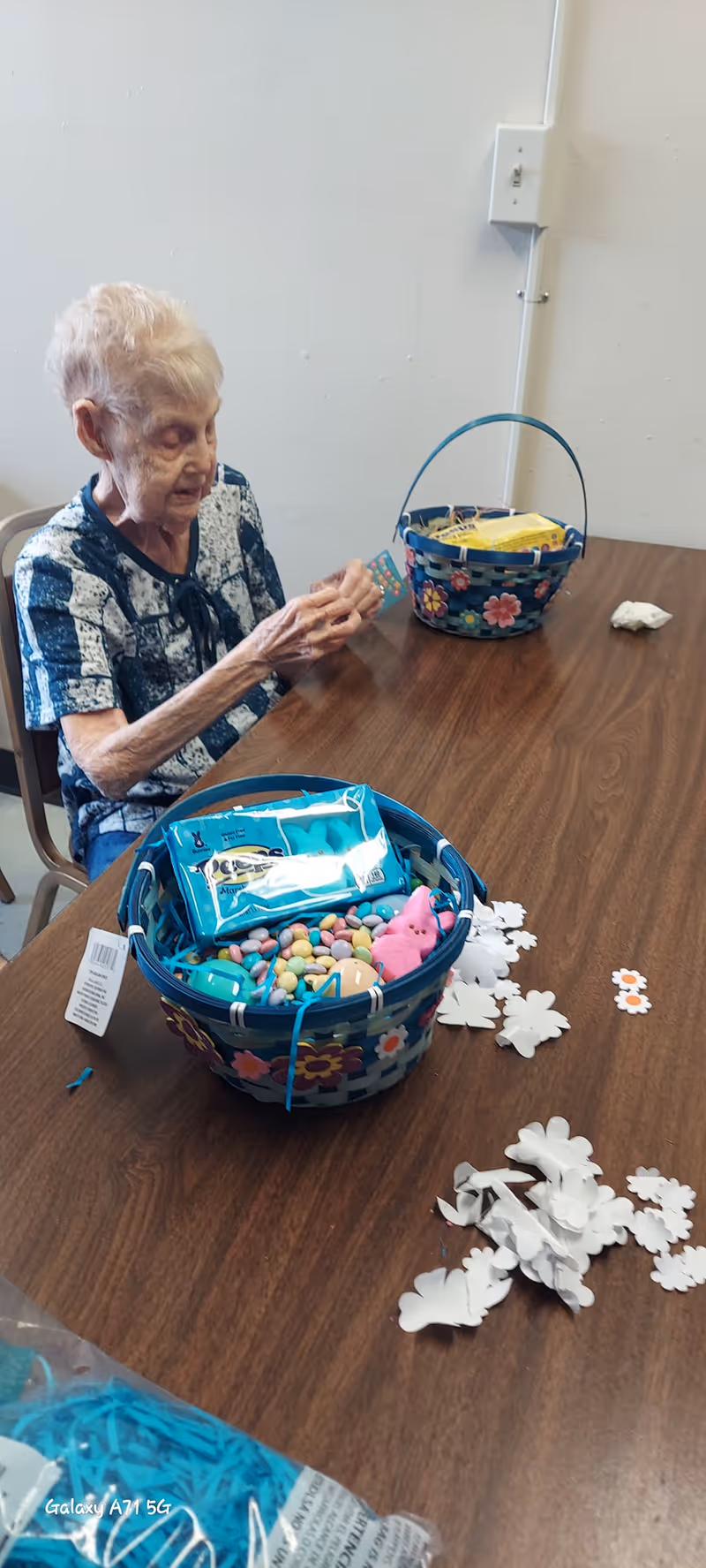 An elderly woman sitting at a wooden table engaged in an activity. On the table are two colorful baskets filled with candy and small items, along with scattered white paper cutouts shaped like flowers and butterflies. The woman appears focused on the activity in a simple room with a plain wall and electrical outlet visible.