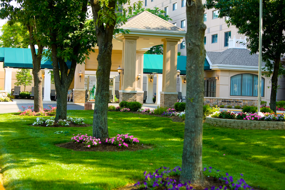 View of the landscaped front entrance of Westminster Village Terre Haute featuring green grass, flower beds with pink, white, and purple flowers, several trees, and a covered driveway with beige pillars and green awnings.