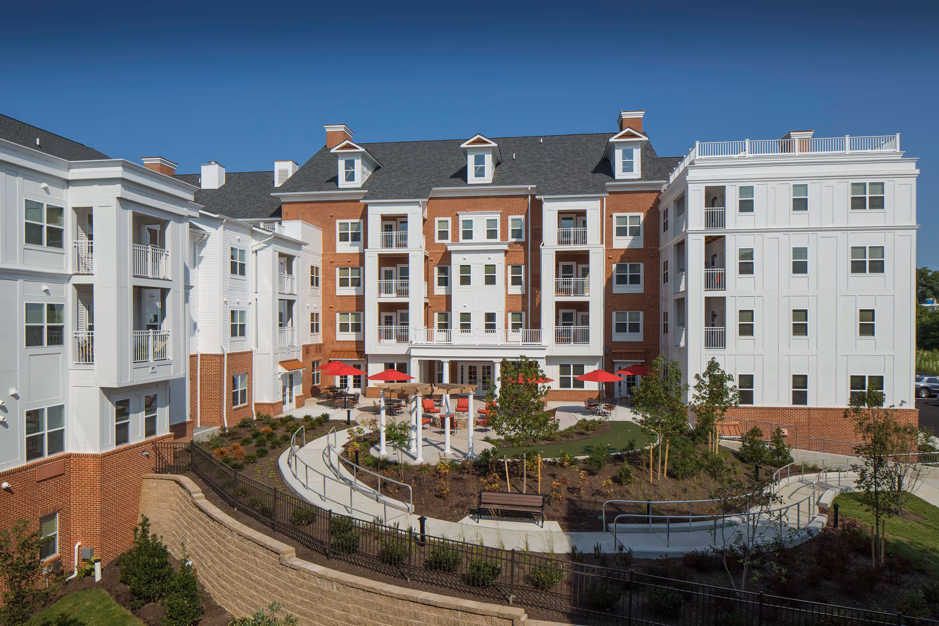 Exterior view of a multi-story senior living facility with a landscaped courtyard featuring walking paths, benches, small trees, and red umbrellas over outdoor seating areas under a clear blue sky.