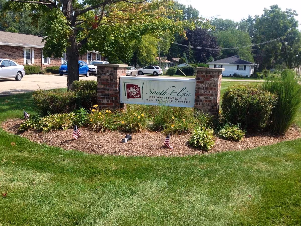 Landscaped entrance sign for South Elgin Rehabilitation & Health Care Center between brick pillars on a lawn with small American flags and parked cars in the background.