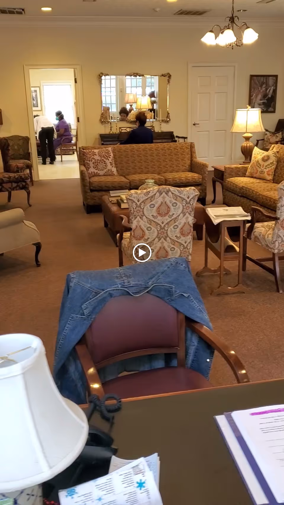 A cozy living room area with patterned armchairs, a sofa, and a coffee table. A denim jacket is draped over a chair in the foreground. In the background, a person is seated at a piano with a large mirror above it, and another person is seen in an adjacent room. The room is warmly lit with table lamps and a ceiling light fixture.