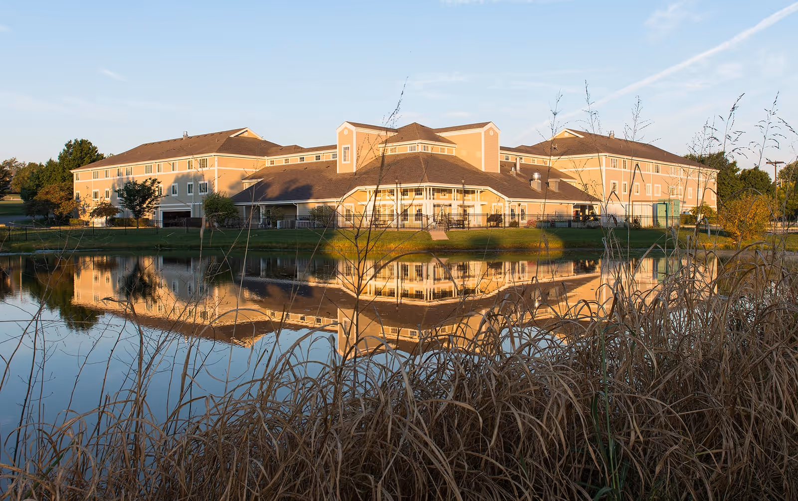 A large beige senior living facility building with multiple windows and a sloped roof, reflected in a calm pond in the foreground. Tall grasses and trees surround the pond, and the sky is clear with soft sunlight illuminating the scene.