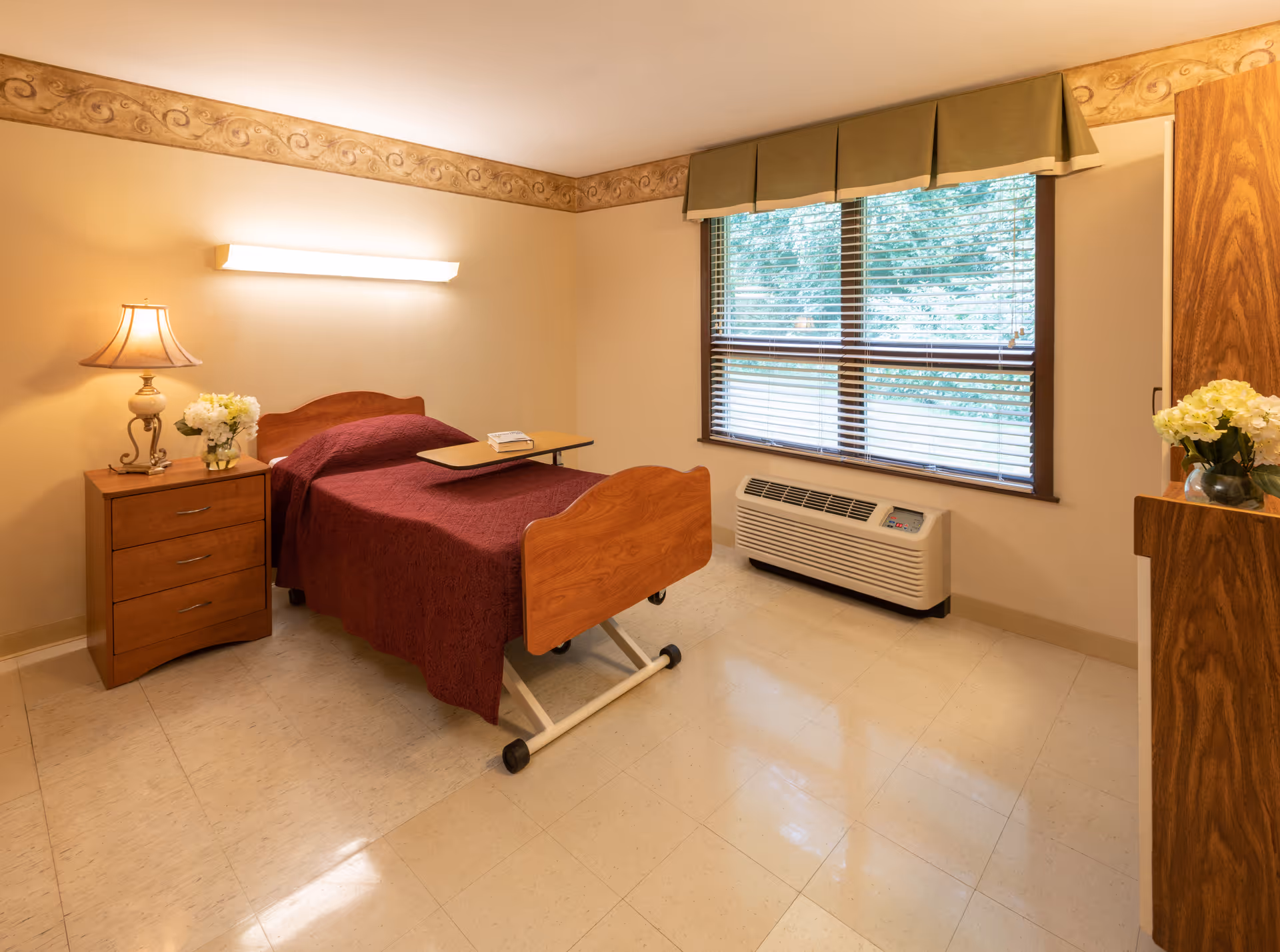 A simple, clean bedroom in a senior living facility featuring a single wooden bed with a maroon bedspread, a wooden nightstand with a lamp and a vase of flowers, a window with blinds and a green valance, and a heating/cooling unit below the window.
