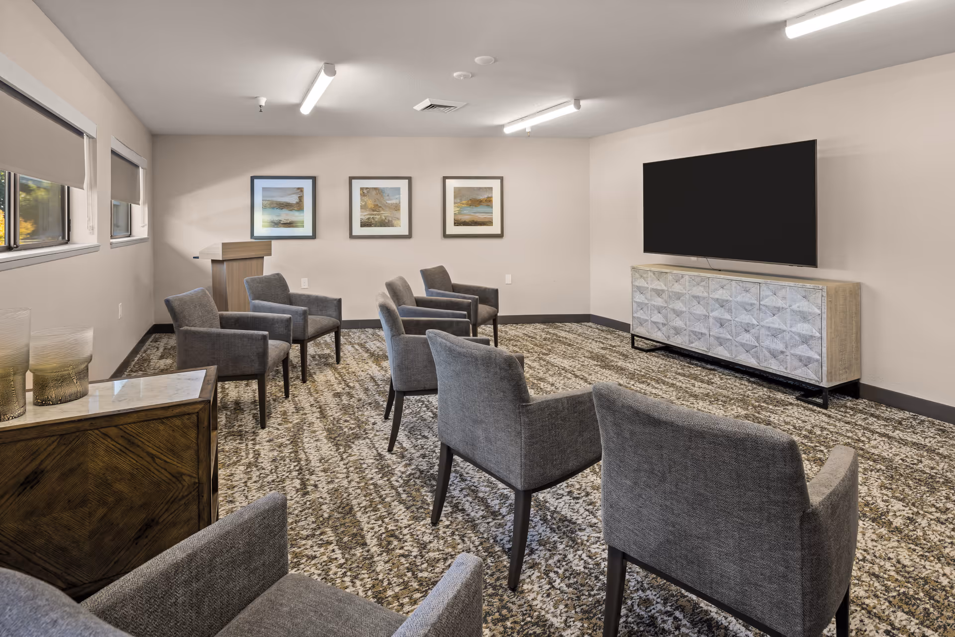 A small meeting or media room with several gray upholstered chairs arranged in rows facing a large flat-screen TV mounted on the wall above a modern cabinet. The room has a patterned carpet, three framed landscape pictures on the wall, and windows with blinds on the left side.