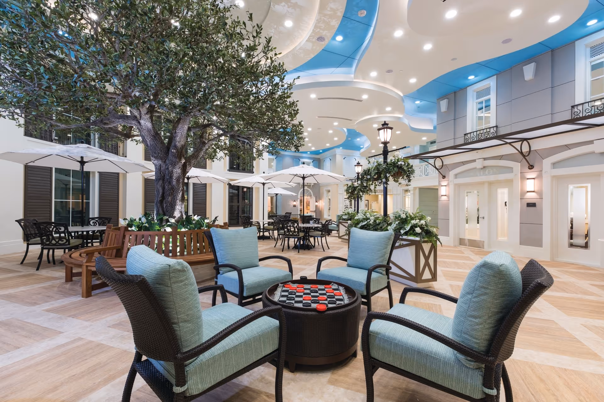 Indoor courtyard area with comfortable seating including four cushioned chairs around a round table with a checkers game. The space features a large tree in the center, several tables with umbrellas, decorative street lamps, and a high ceiling with blue and white curved design elements and recessed lighting.