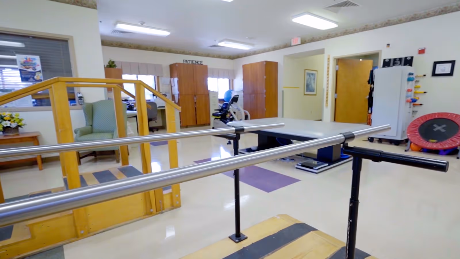 Rehabilitation therapy room with parallel walking bars, a ramp, exercise equipment, chairs and storage cabinets.